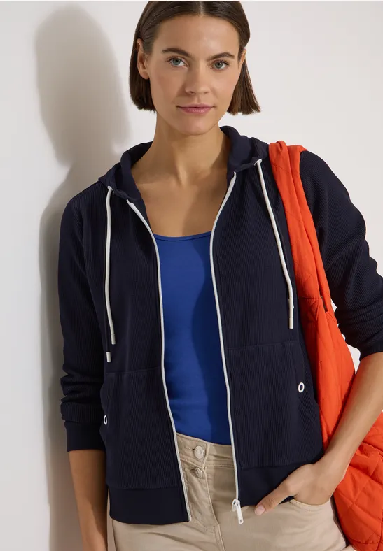 Person trägt dunkelblauen Hoodie, blaues Top und helle Hose, hält rote Tasche über der Schulter.