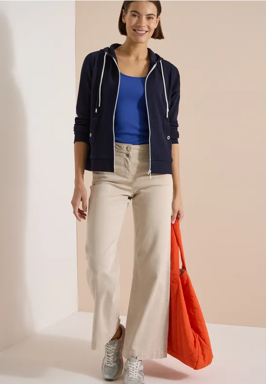 Frau trägt beige weite Hose, blaues Top, dunkelblaue Kapuzenjacke, graue Sneaker und orange Tasche.