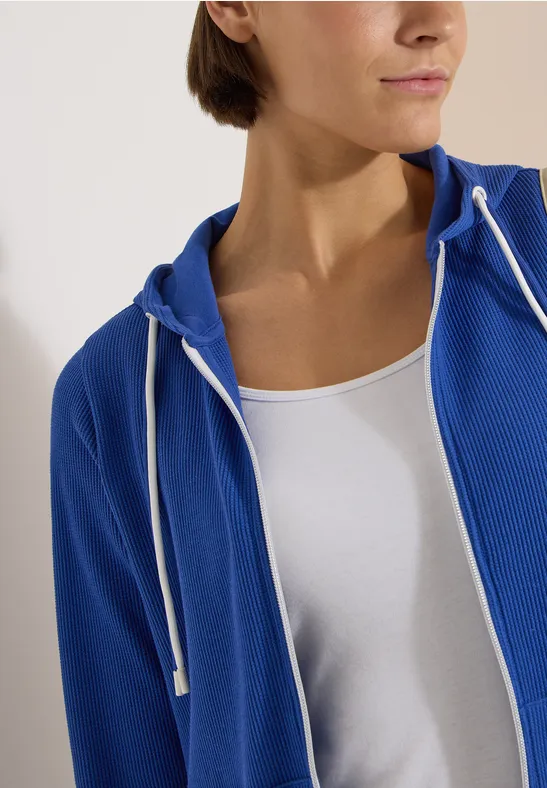 Teilansicht einer Frau mit weißem Top und blauer Kapuzenjacke, Fokus auf den Oberkörper