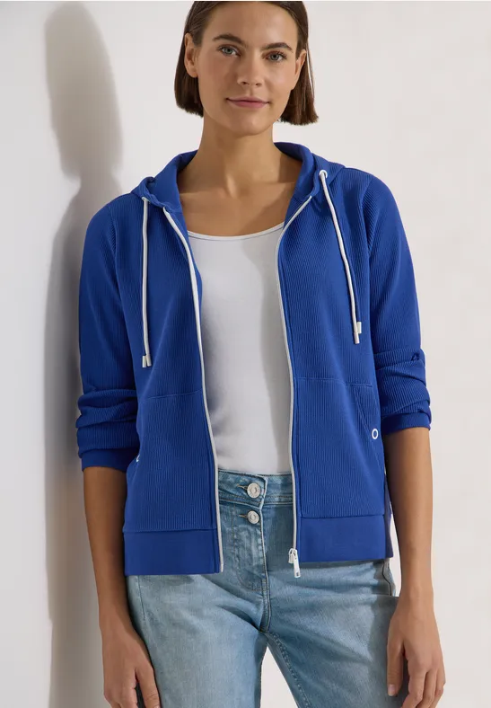 Eine Person trägt blaue Kapuzenjacke, weißes Shirt und Jeans vor hellem Hintergrund.