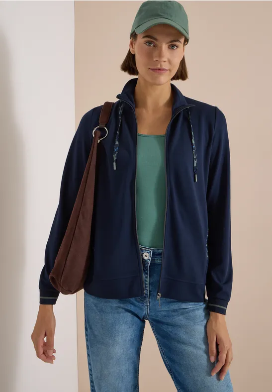 Frau trägt Jeans, grünes Top, blaue Kapuzenjacke, grünes Cap und braune Schultertasche.