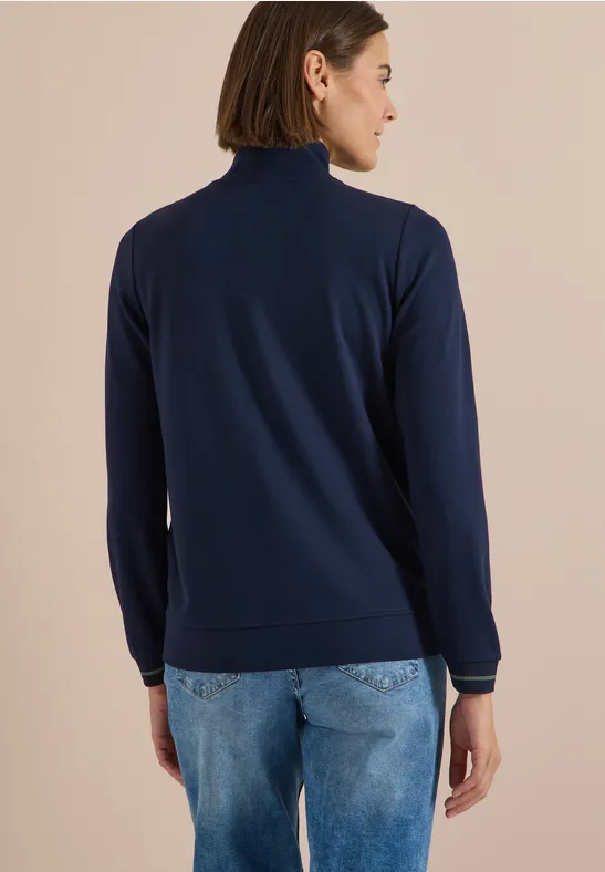 Rückansicht einer Person in blauem Rollkragenpullover und Jeans vor neutralem Hintergrund.