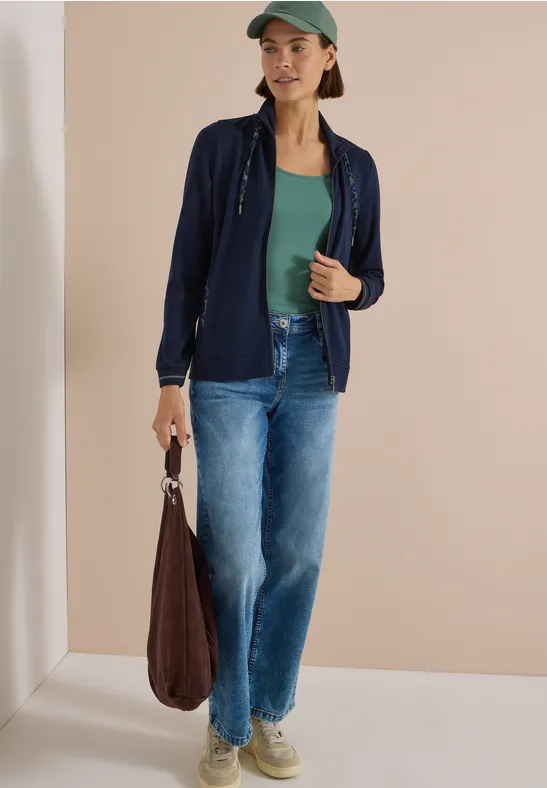 Eine Frau trägt grünes Top, marineblaue Jacke, blaue Jeans, Cap und hält eine braune Tasche.