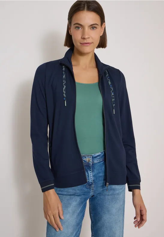 Eine Frau mit kurzen braunen Haaren, dunkelblauer Kapuzenjacke, grünem Top und Jeans steht vor hellem Hintergrund.