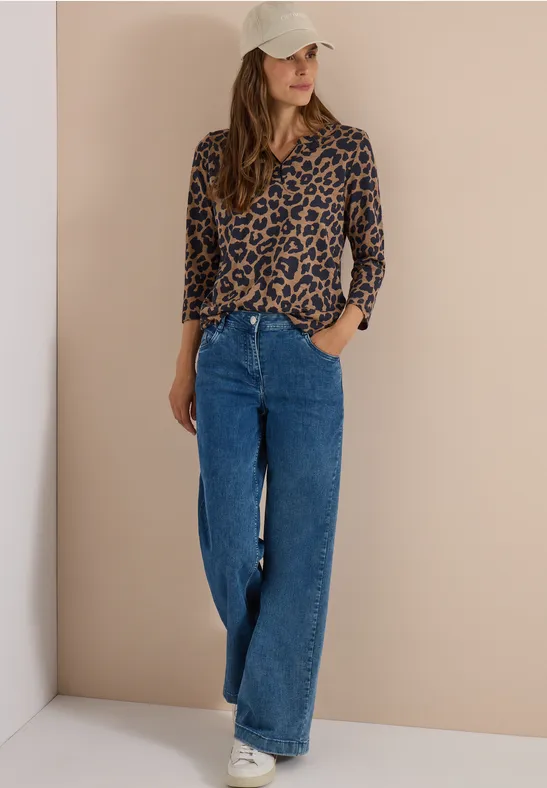 Frau mit Leopardenmuster-Bluse, weiten Jeans, Baseballcap und Sneakern steht vor beiger Wand.