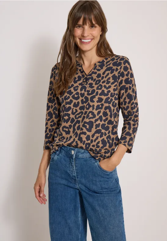 Frau trägt eine Leo-Bluse und blaue Jeans, lächelt entspannt mit einer Hand in der Hosentasche.