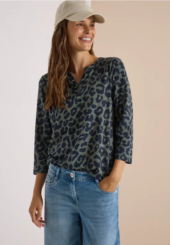 Frau in Baseballcap, grüner Leoparden-Longsleeve und Jeans lächelt mit lockerer Pose.
