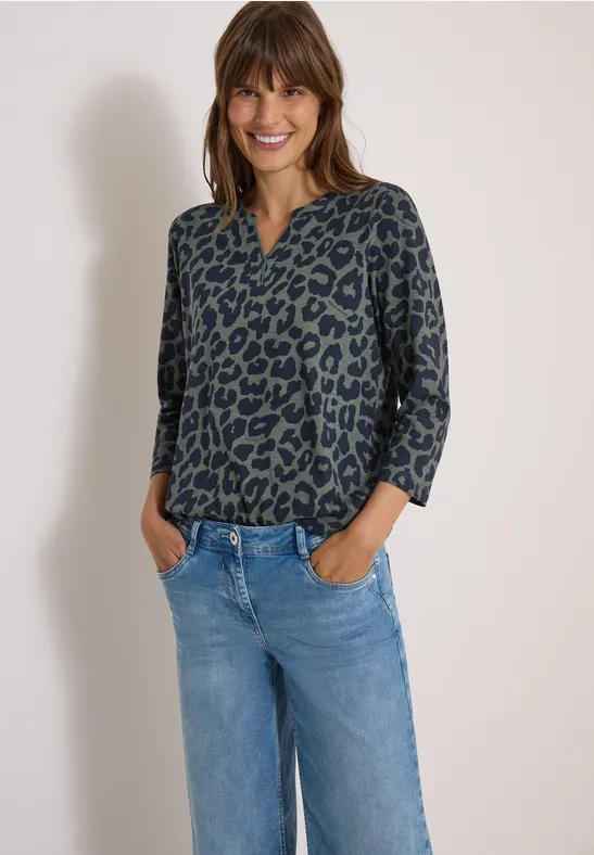 Frau steht lächelnd in legerer Bluse mit Leopardenmuster und blauer Jeans, Hände in den Taschen.