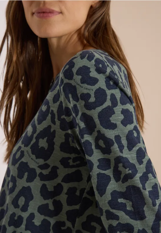 Eine Frau mit braunen Haaren trägt ein blau-graues Shirt mit schwarzem Leopardenmuster.