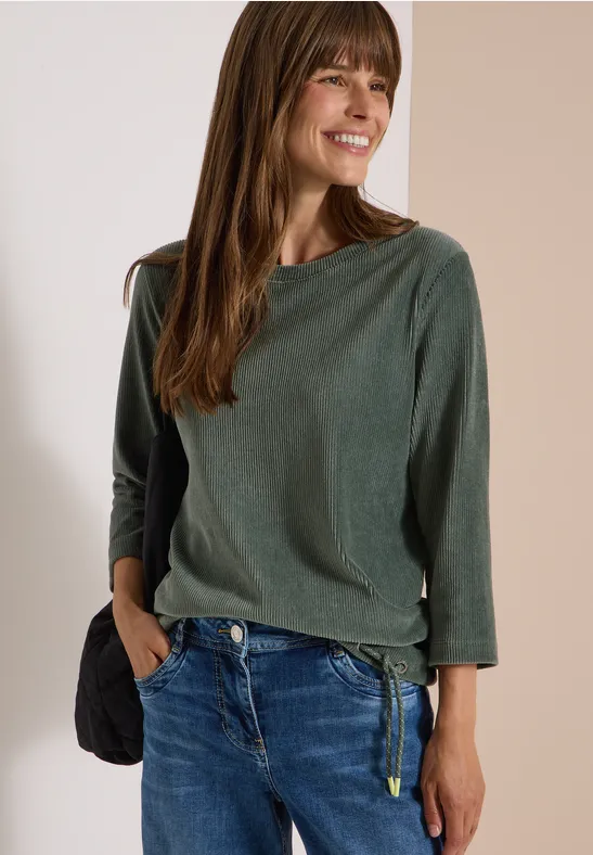 Femme souriante en haut vert à manches longues et jean bleu, main dans la poche, posant devant un mur clair.