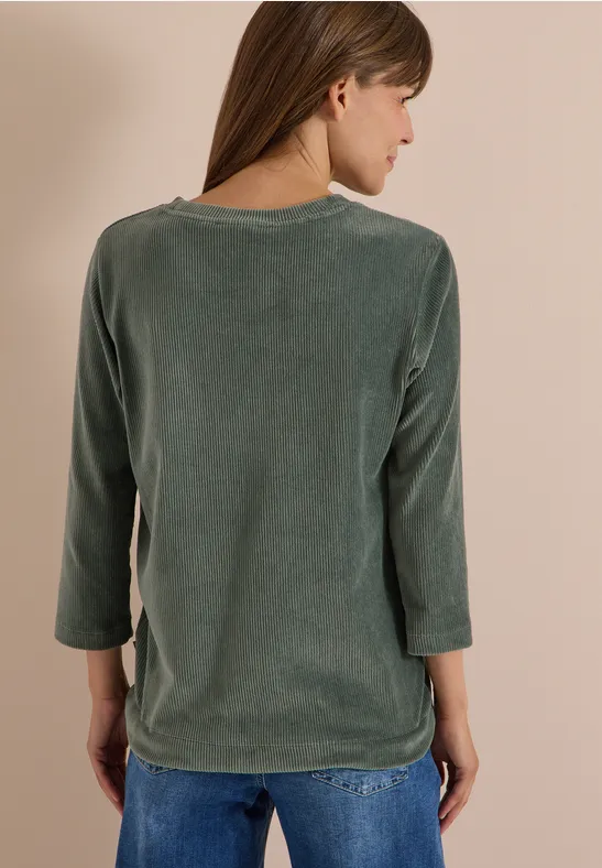 Une femme vue de dos porte un t-shirt vert à manches longues et un jean bleu.