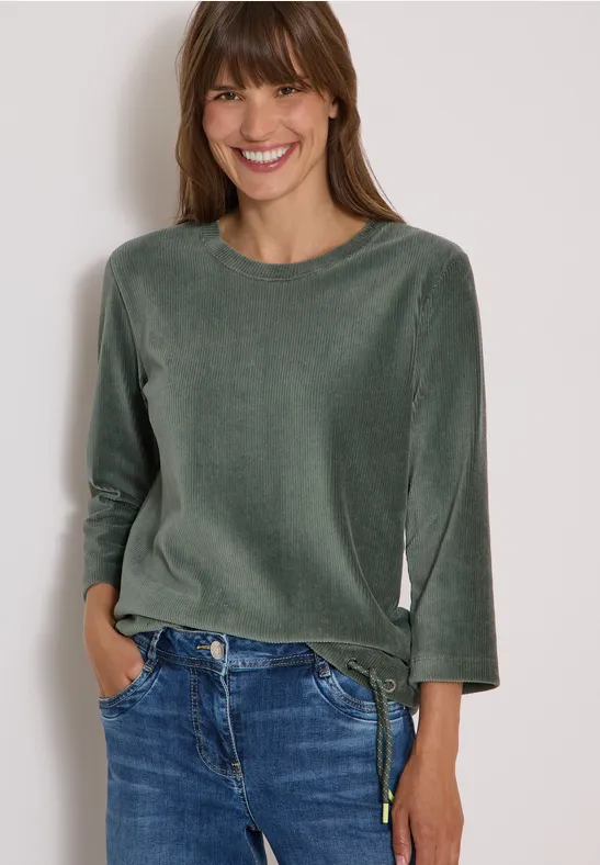 Une femme souriante porte un haut vert en velours et un jean bleu, les mains dans les poches.
