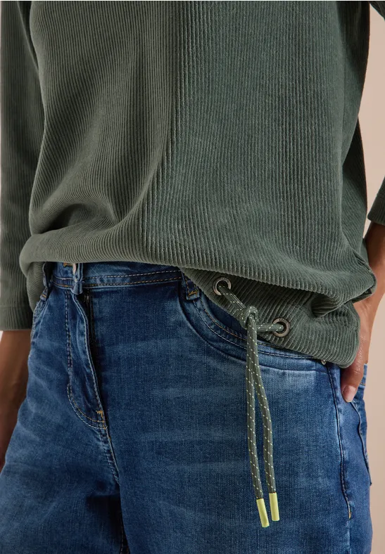 Jean à cordon vert et pull côtelé vert, main dans la poche.