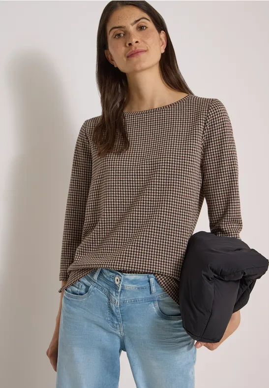 Femme en haut à carreaux et jeans, tenant une doudoune noire sous le bras.