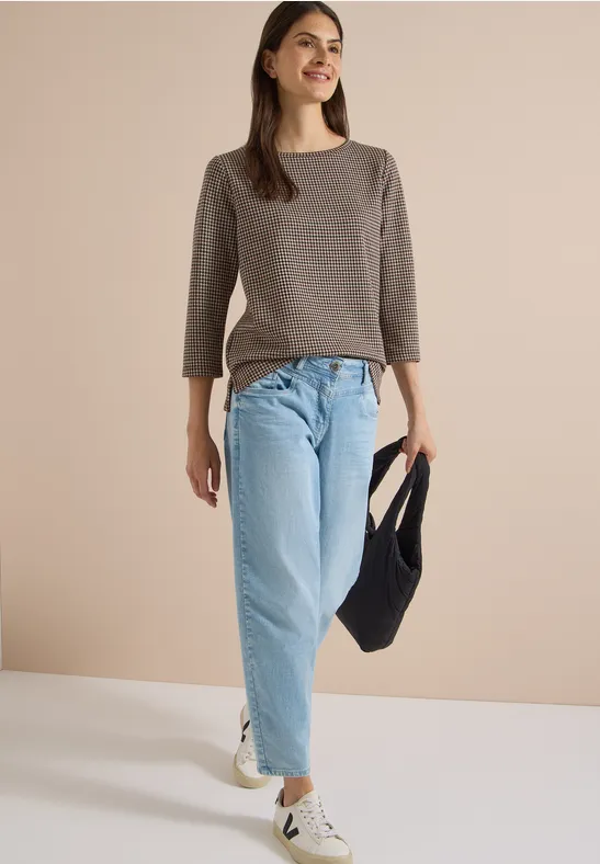 Femme en haut gris à manches 3/4, jean bleu clair ample, baskets blanches et sac noir sur fond clair.