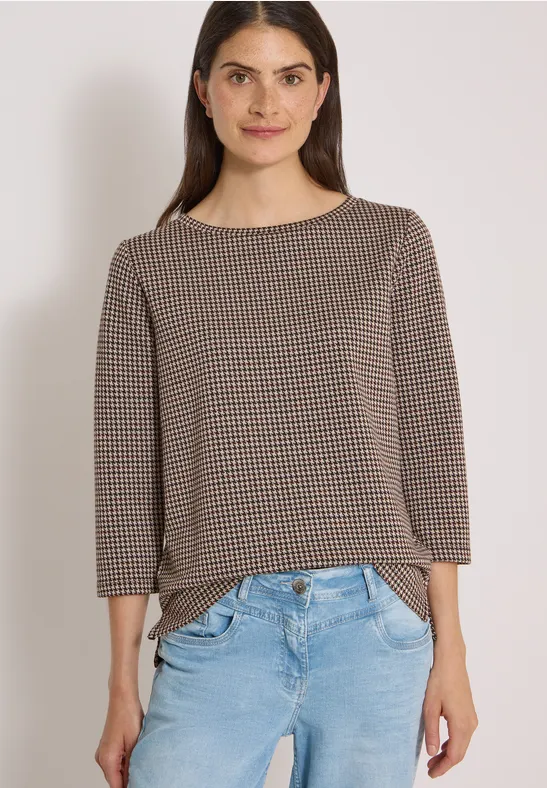 Femme souriante portant un pull à carreaux marron-beige et un jean bleu clair sur fond clair.