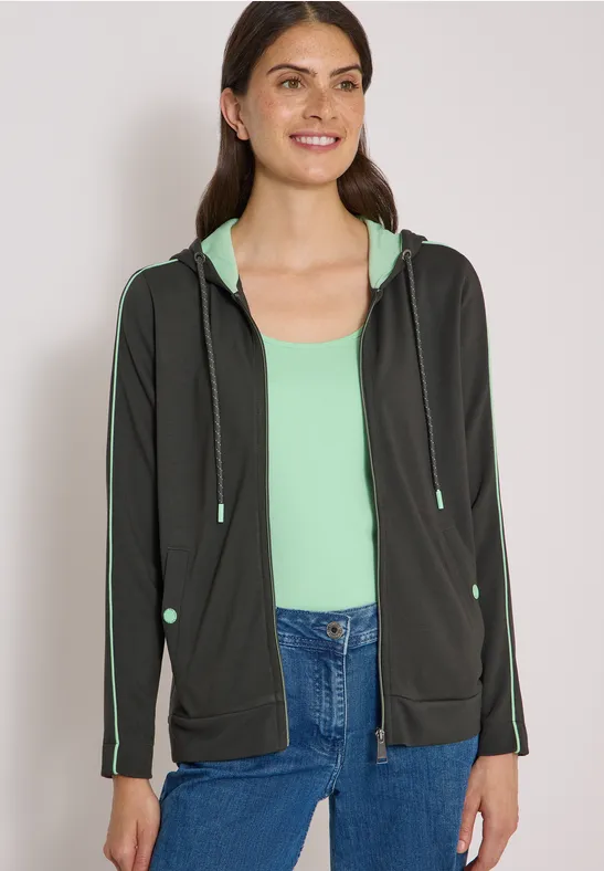 Frau in schwarzer Kapuzenjacke und mintgrünem Top, lächelnd und blickt geradeaus.