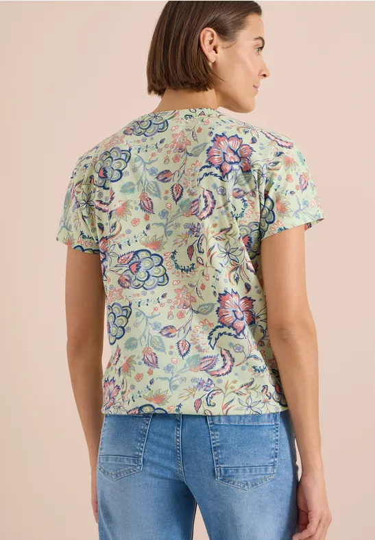 Femme de dos portant un haut fleuri à manches courtes et un jean sur fond neutre.