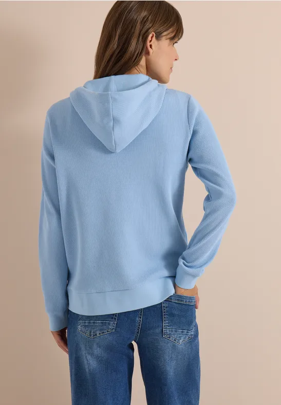 Frau von hinten in hellblauer Kapuzenjacke und blauen Jeans vor neutralem Hintergrund.