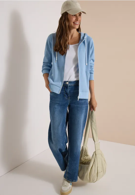 Frau trägt weißes Top, hellblauen Hoodie, weite Jeans, Cap und große Tasche.
