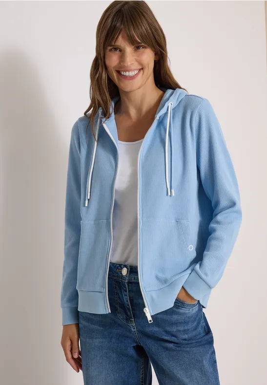 Frau mit hellblauem Kapuzen-Sweatshirt, weißem T-Shirt und Jeans lächelt vor weißem Hintergrund.