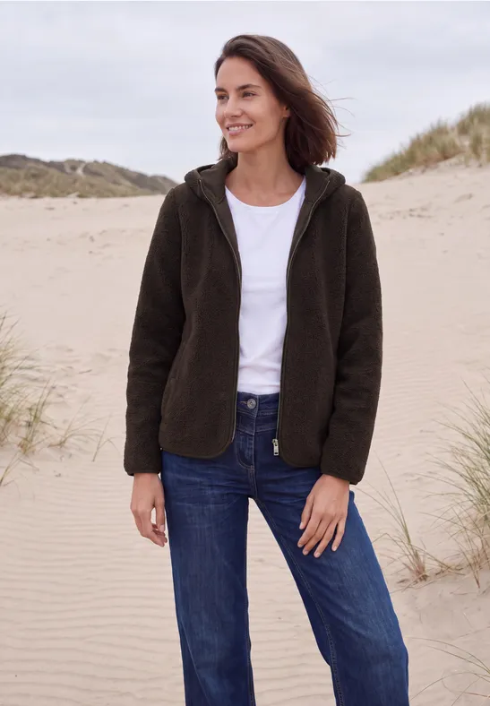 Frau in dunkelgrüner Fleecejacke, weißem T-Shirt und blauen Jeans steht lächelnd am Dünenstrand