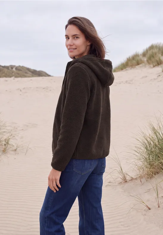 Frau in brauner Kapuzenjacke und blauer Hose auf sandiger Dünenlandschaft, blickt über die Schulter.