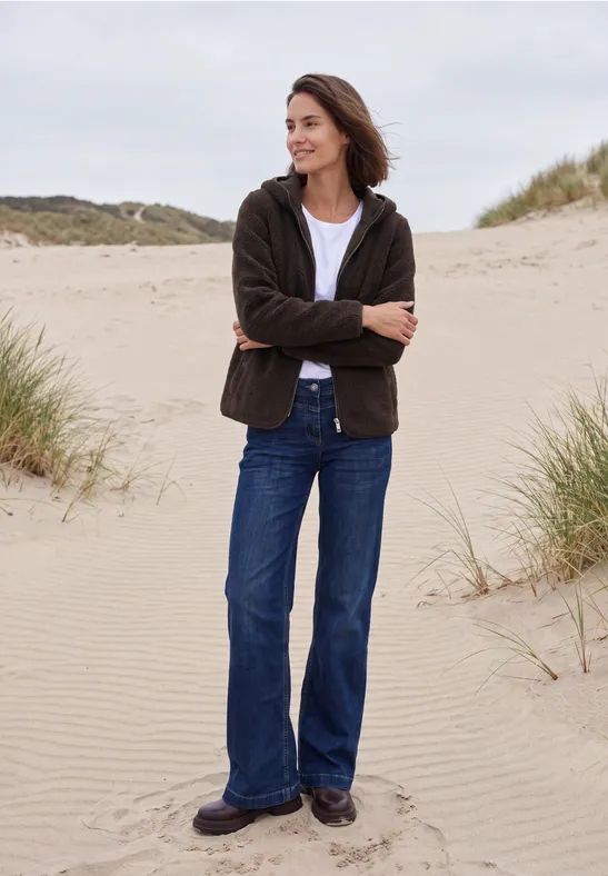 Frau in Jeans und Jacke steht entspannt mit verschränkten Armen auf Dünen am Strand.