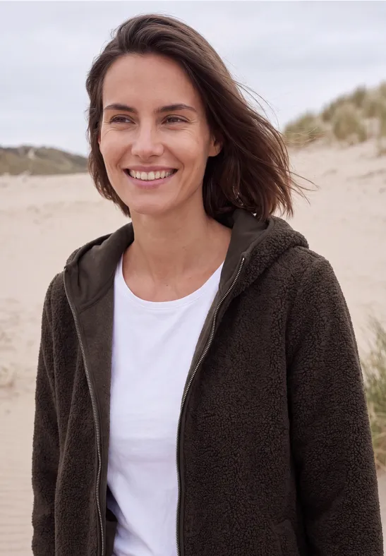 Eine lächelnde Frau in einem Fleece-Pullover steht am Strand mit Dünen im Hintergrund.