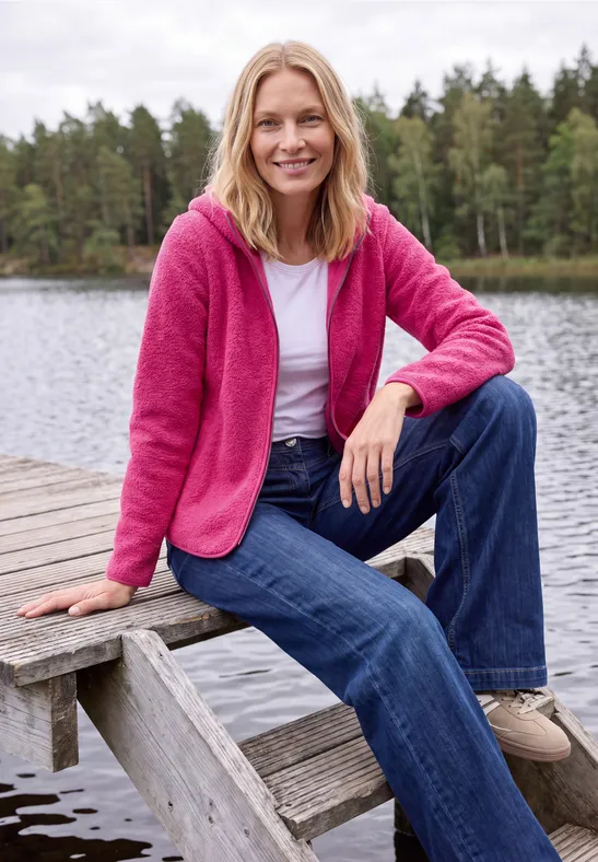 Eine Frau in pinkem Pullover und weißem Shirt sitzt auf einem Steg vor einem Waldsee.