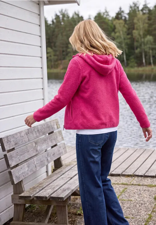 Eine Person im pinken Hoodie und Jeans steht am Steg am See mit Holzbank und Wald im Hintergrund.