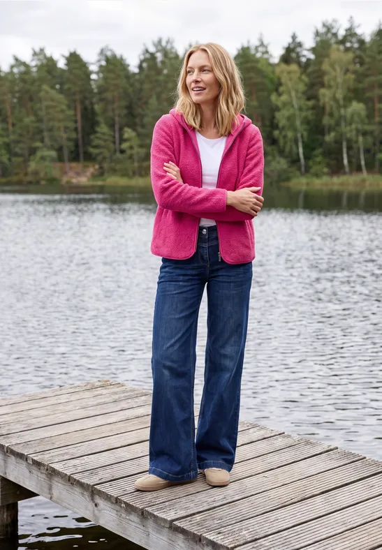 Eine Frau in pinkem Pullover und Jeans steht entspannt auf einem Holzsteg am See, Wald im Hintergrund.