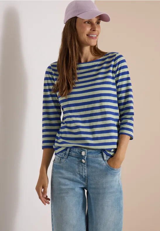 Femme en marinière, jean et casquette rose, souriante, une main dans la poche.
