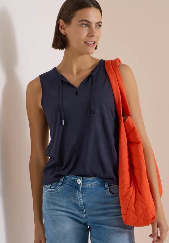 Frau in ärmellosem dunkelblauem Top und Jeans, trägt eine orange Steppjacke über der Schulter.