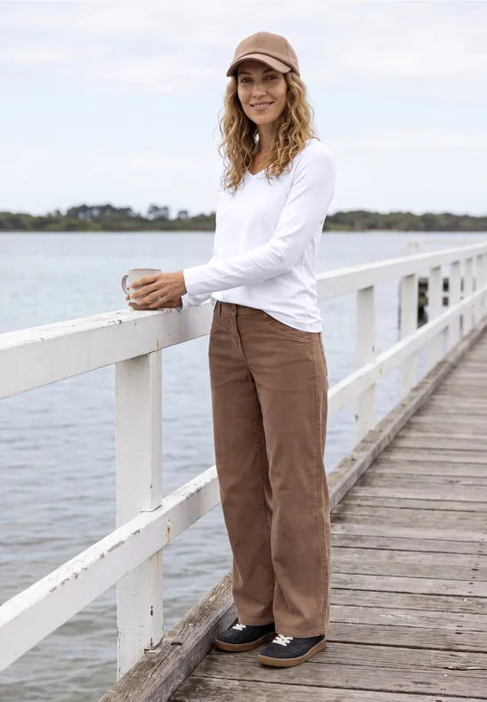 Frau in weißem Longsleeve, braunen Hosen und Cap steht entspannt auf einem Steg am Wasser.