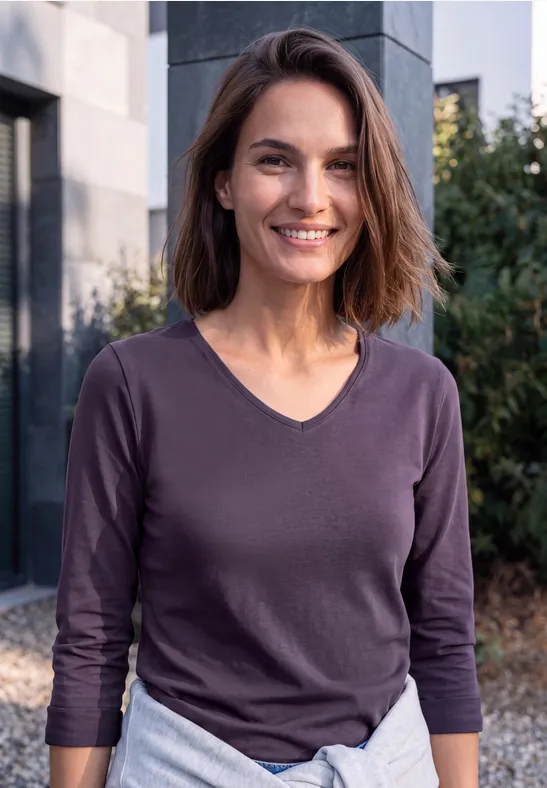Une femme aux cheveux bruns souriante, vêtue d'un haut violet, devant un bâtiment moderne.