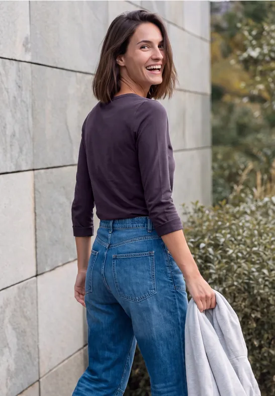 Femme souriante en jean et haut à manches longues, tenant une veste et regardant par-dessus son épaule.