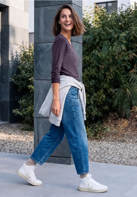 Femme en jean, baskets et pull, souriante, marche devant un bâtiment et des buissons, pull en plus noué à la taille.