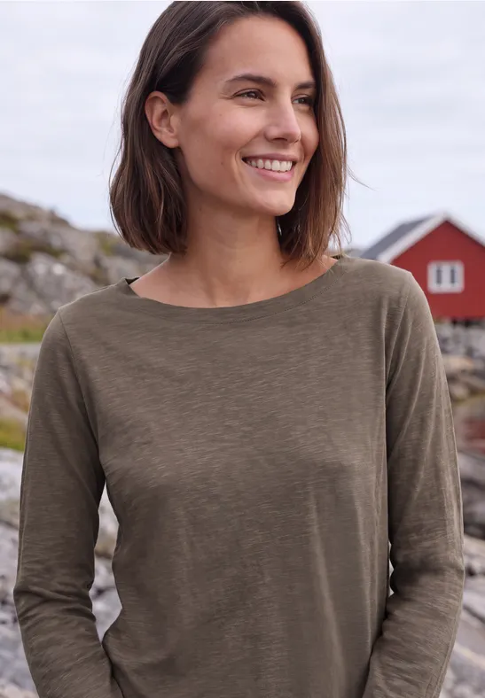 Lächelnde Frau in olivgrünem Pullover, im Hintergrund Felsen und rote Häuschen an der Küste.