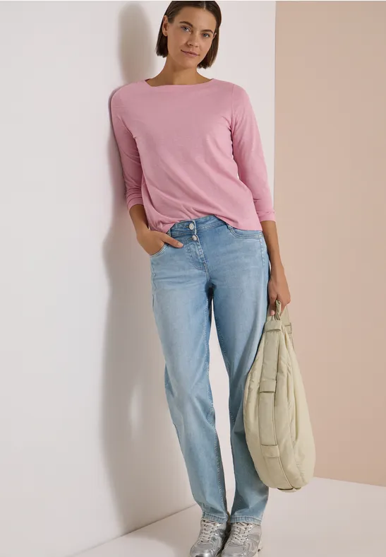 Une femme souriante en haut rose et jean bleu appuyée contre un mur, tenant un grand sac beige.
