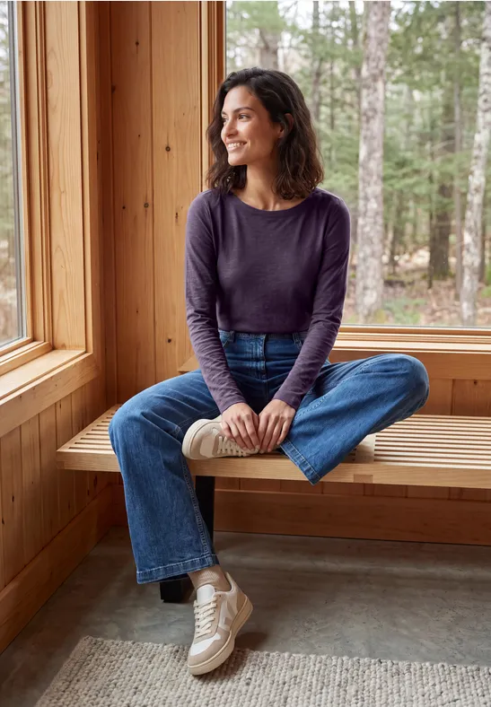 Une femme souriante en jean et pull à manches longues est assise sur un banc en bois près d'une fenêtre dans un chalet.