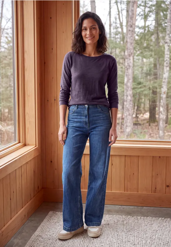 Femme debout dans une pièce en bois près d'une fenêtre, porte t-shirt manches longues, jean large et baskets.