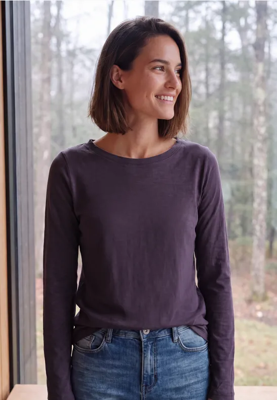 Une femme souriante aux cheveux bruns, en haut violet et jean, regarde par une fenêtre sur la forêt