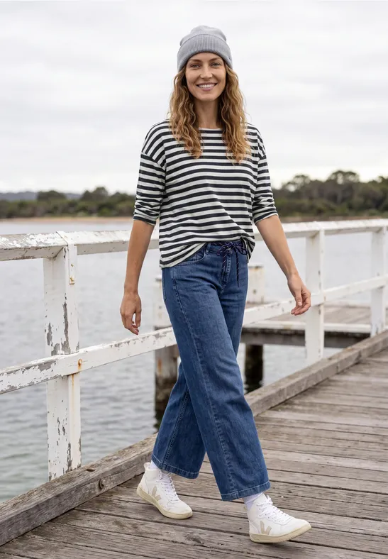 Une femme en jean, marinière, bonnet et baskets marche en souriant sur une jetée au bord de l’eau.