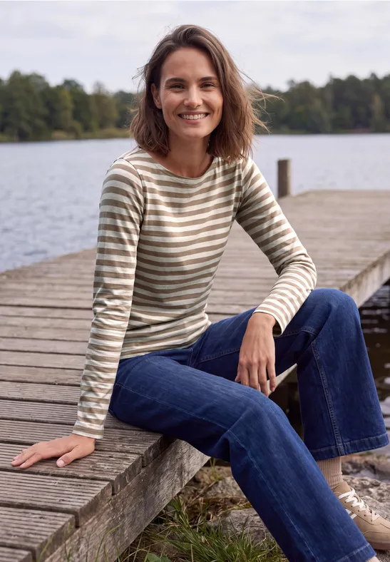 Une femme souriante assise sur un ponton en bois au bord d’un lac, vêtue d’un t-shirt rayé et d’un jean.