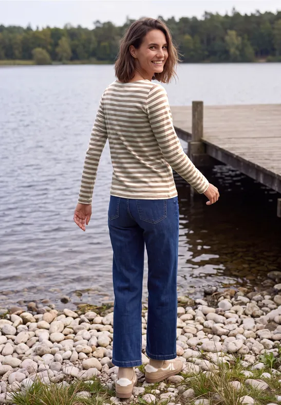 Une femme souriante en pull rayé se promène au bord d'un lac calme, près d'une jetée en bois.