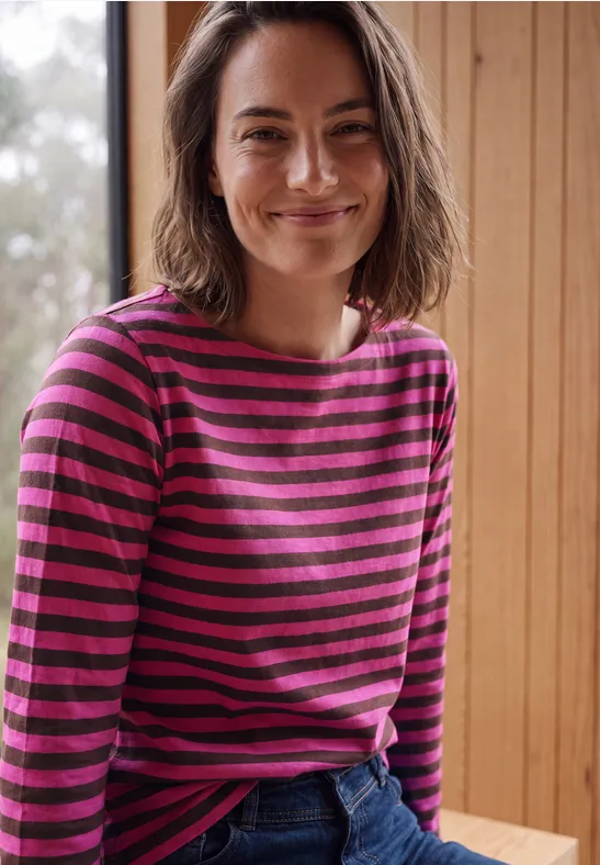 Lächelnde Frau in gestreiftem pink-lila Pullover vor hellem Holzhintergrund.