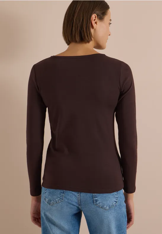 Frau von hinten mit kurzen Haaren, dunkelbraunem Langarmshirt und blauer Jeans vor beigem Hintergrund.