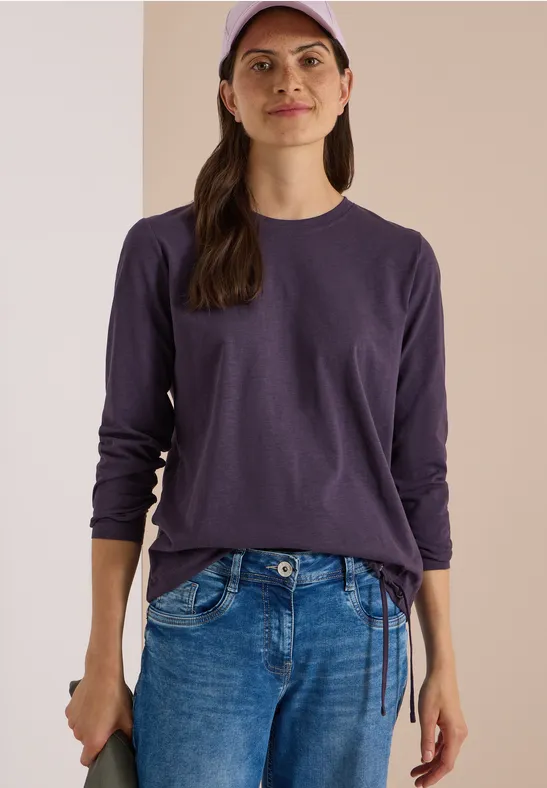 Femme en t-shirt à manches longues violet, jean bleu et casquette sur fond simple.
