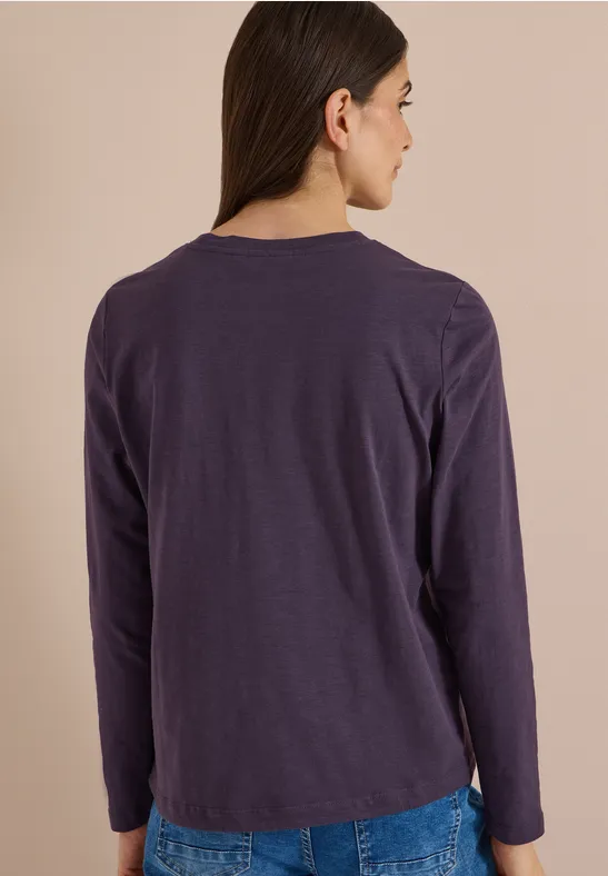 Femme de dos portant un haut uni violet à manches longues devant un fond clair.
